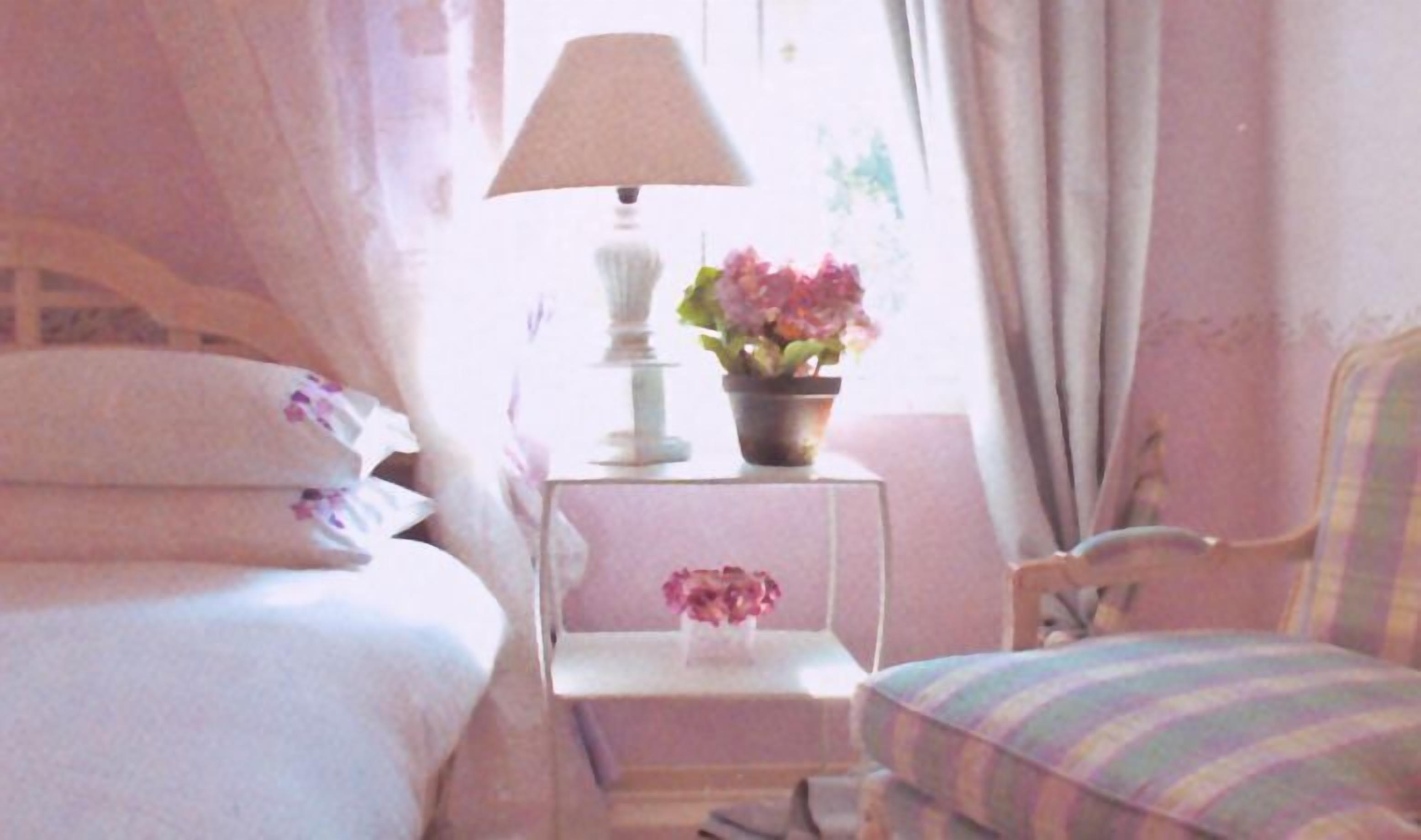 Un dormitori amb un llit, una tauleta de nit amb una làmpada i flors, una cadira i una finestra amb cortines.