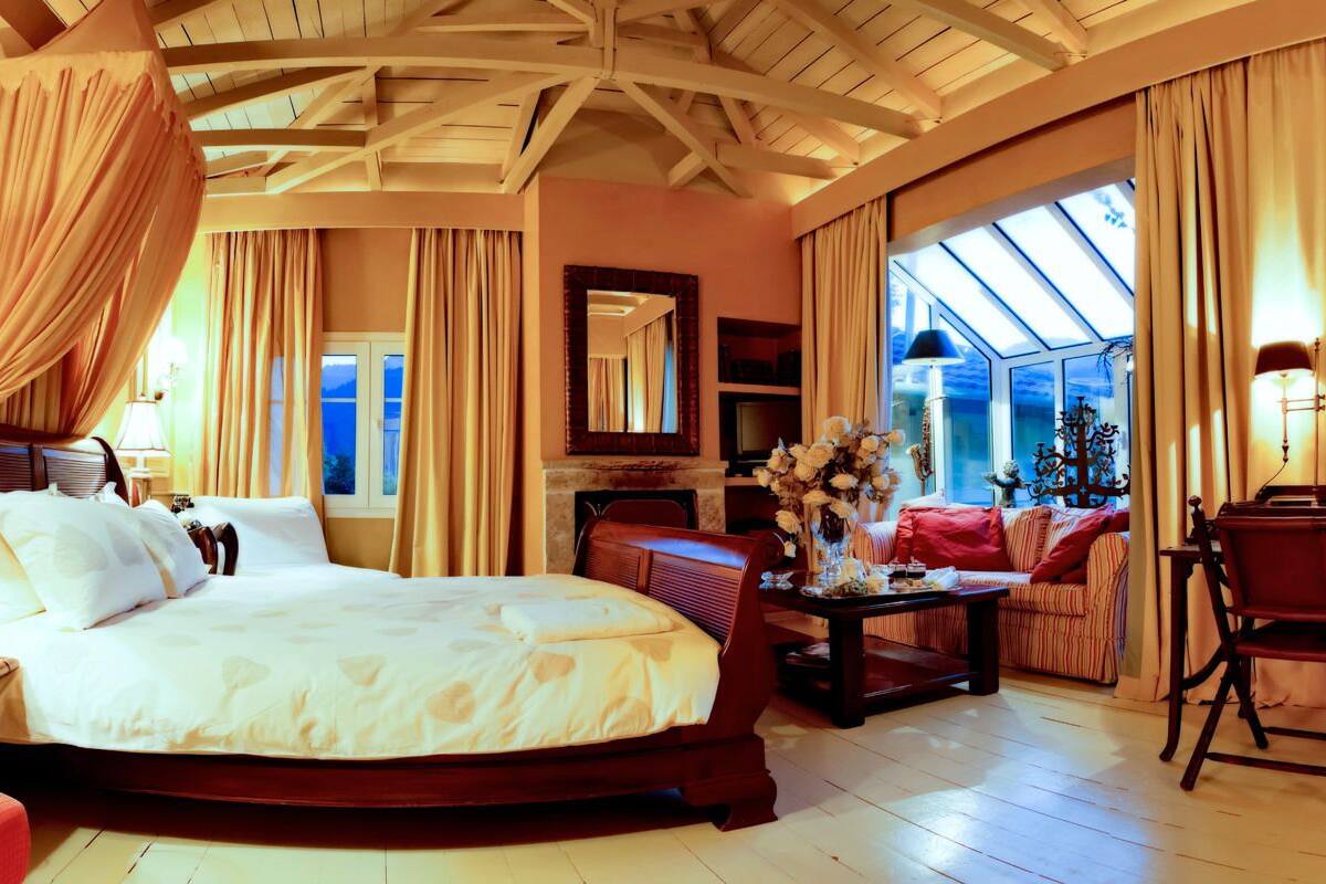 Un dormitorio amplio con una cama grande, una zona de estar con sofá, una mesa de comedor y una araña. Cuenta con un ventanal que proporciona luz natural y vistas al exterior.
