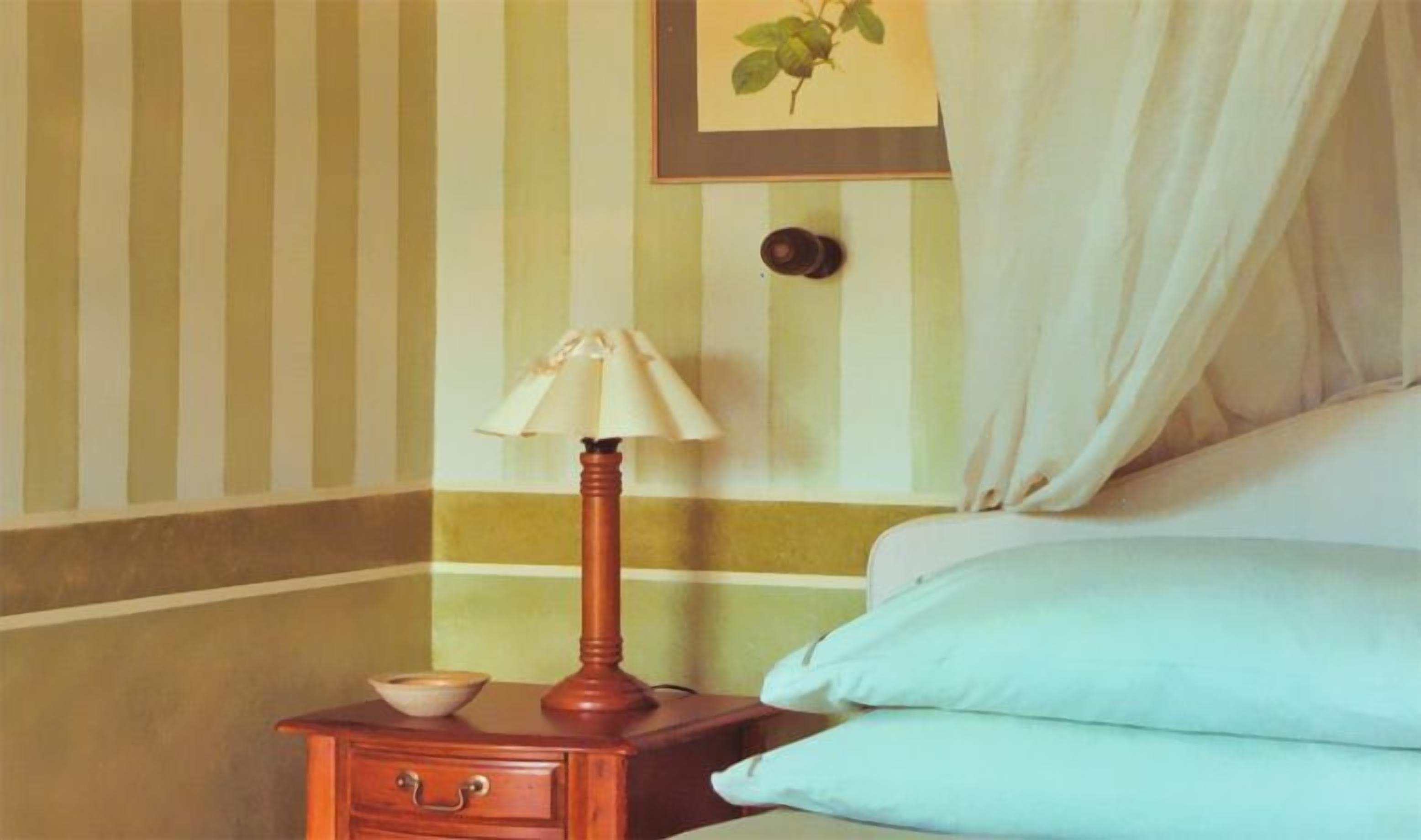 A hotel room with striped wallpaper, a bedside table with a lamp, a bowl, and a bed with light blue pillows.
