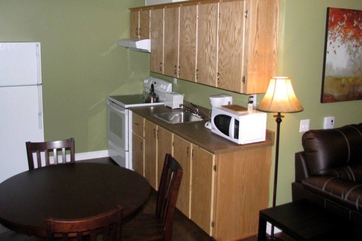 Cocina compacta con gabinetes de madera, un microondas, una estufa y un lavabo. Hay una mesa de comedor con sillas y un sofá en la sala.