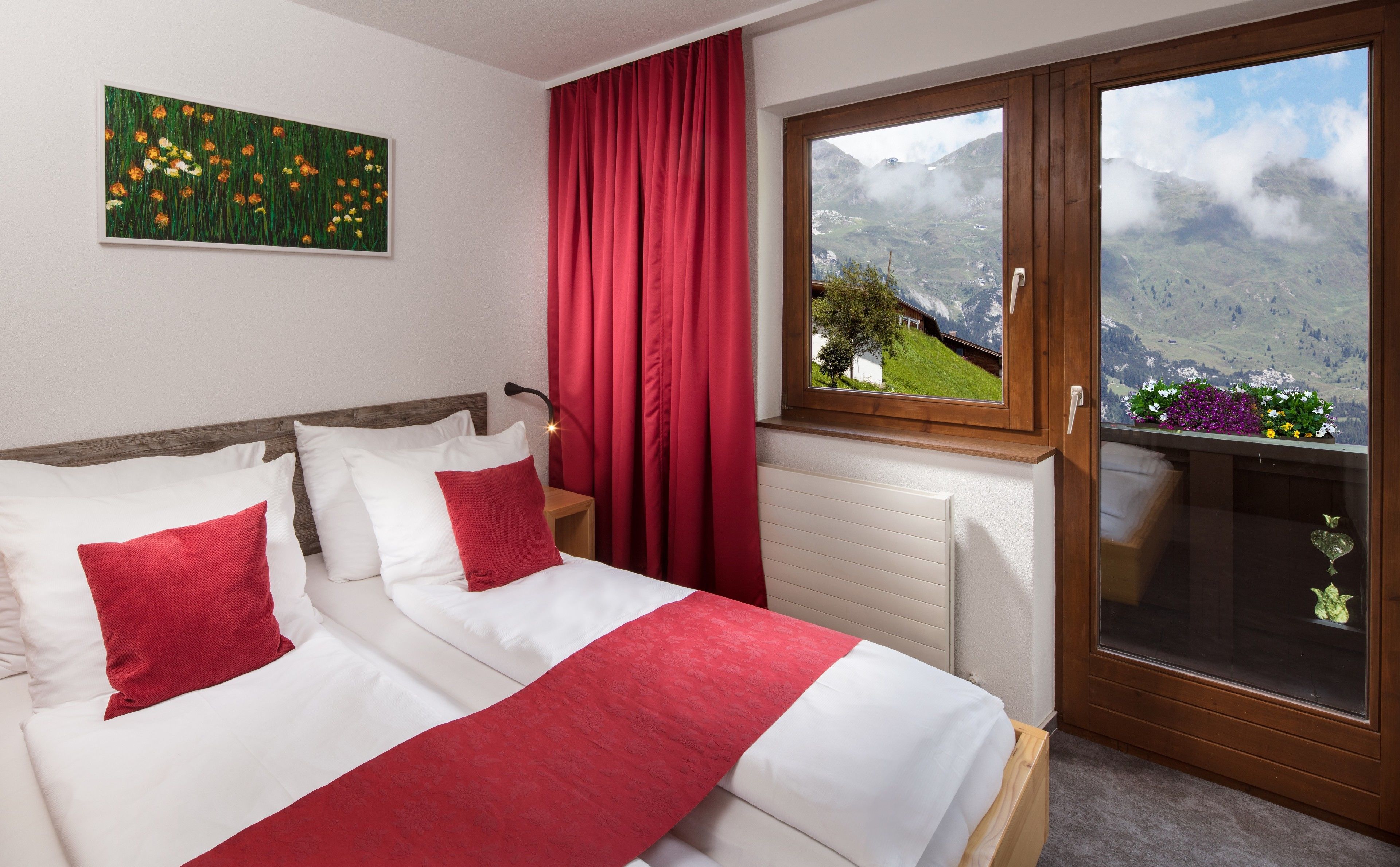 A hotel room with a bed, red curtains, a window with a mountain view, and a painting on the wall.