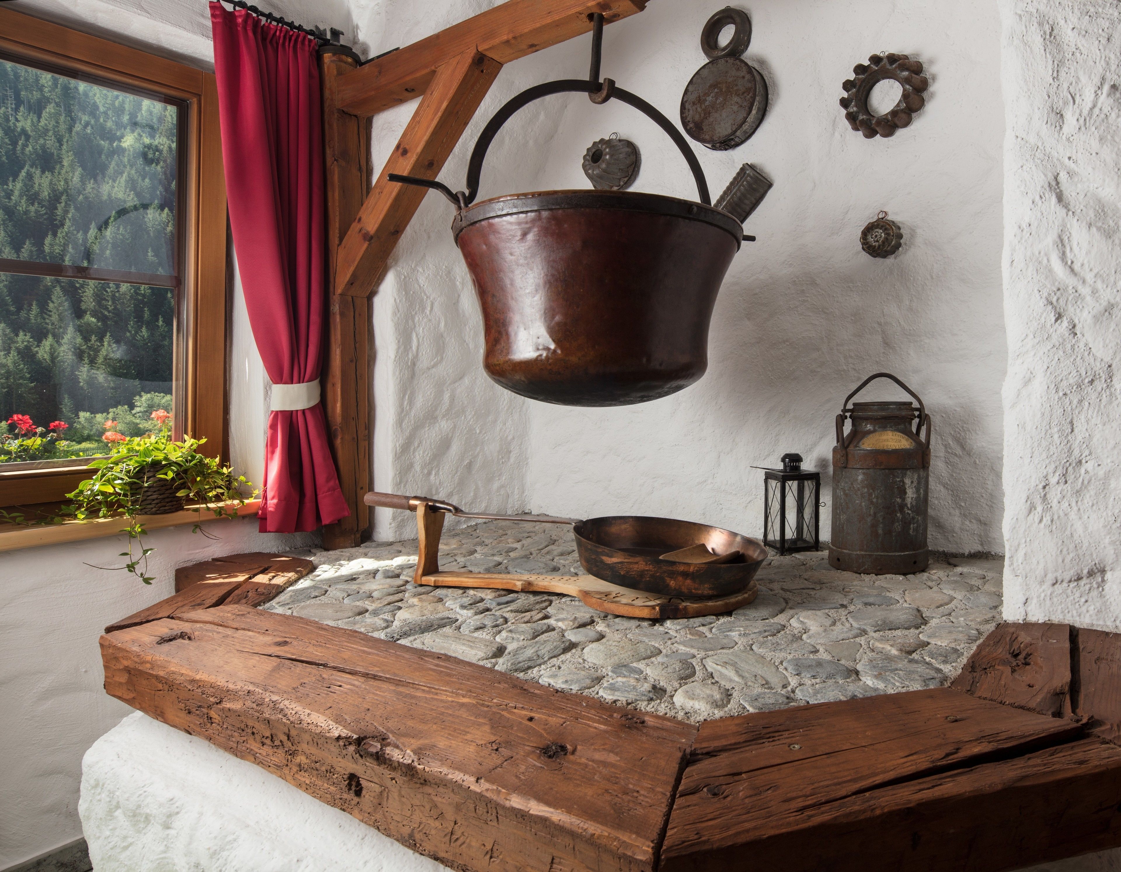 A rustic kitchen with a hanging copper pot, a wooden bench, a lantern, and a window with a red curtain.