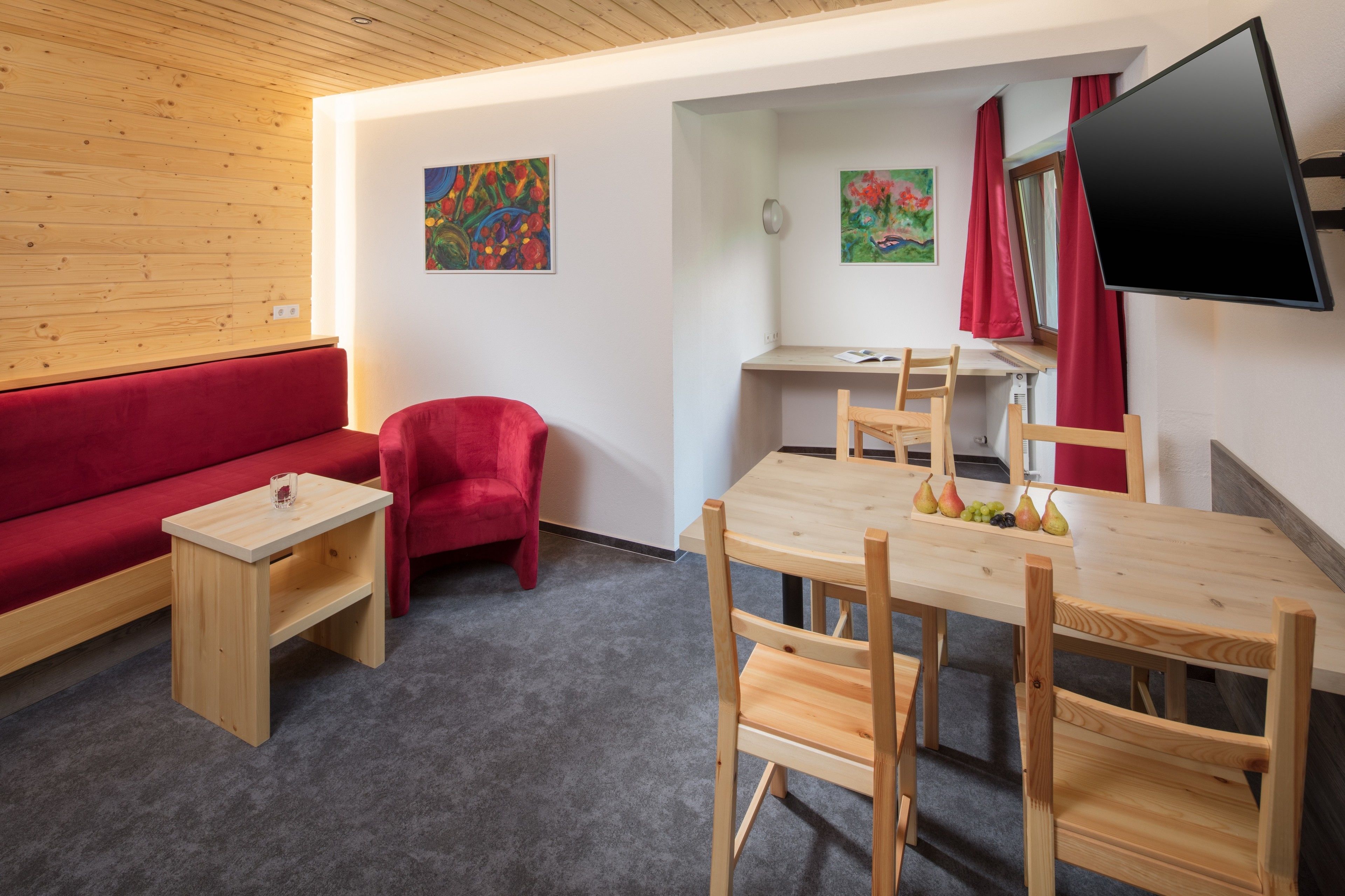 Ein Zimmer mit einer roten Couch, einem kleinen Holztisch, einem Holzbüro mit Stuhl und einem an der Wand montierten Fernseher.