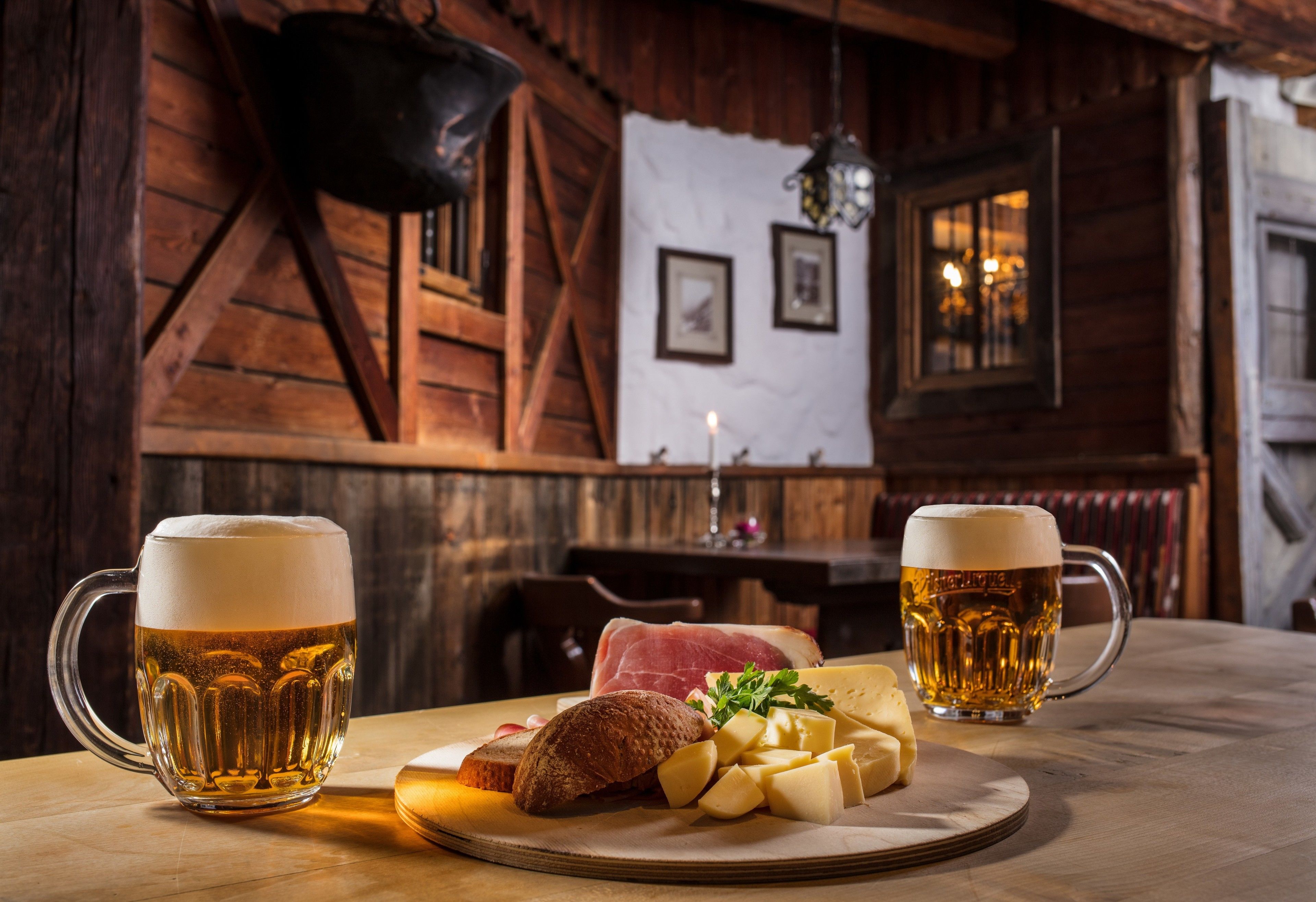 Interior de madera con dos jarras de cerveza, un plato de queso y pan, y una vela sobre una mesa.