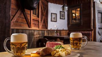 Interior de madera con dos jarras de cerveza, un plato de queso y pan, y una vela sobre una mesa.