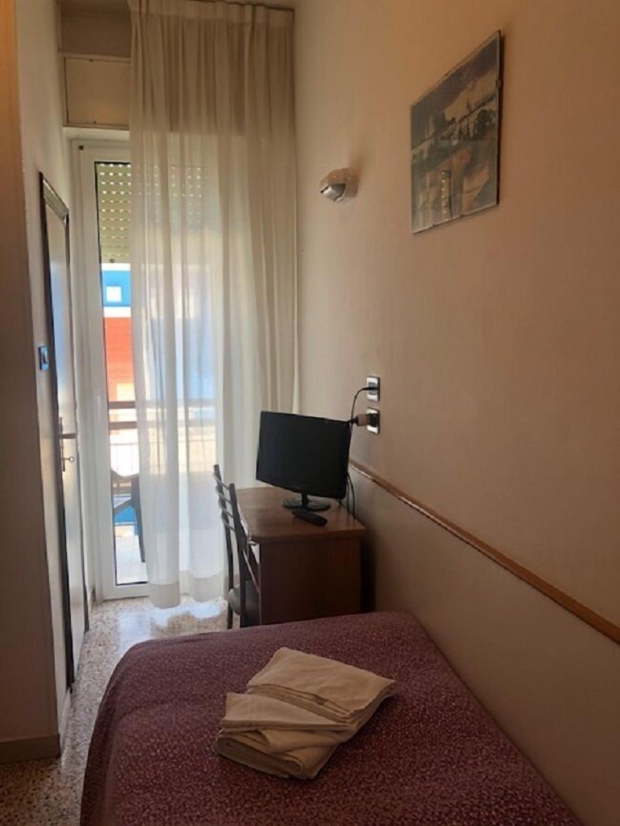 A hotel room with a bed, a desk with a computer, a painting on the wall, and a door leading to a balcony.