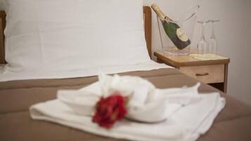 A neatly made bed with a white pillow, a folded white towel, and a red rose. A wooden nightstand beside the bed holds a bottle of champagne and two glasses.