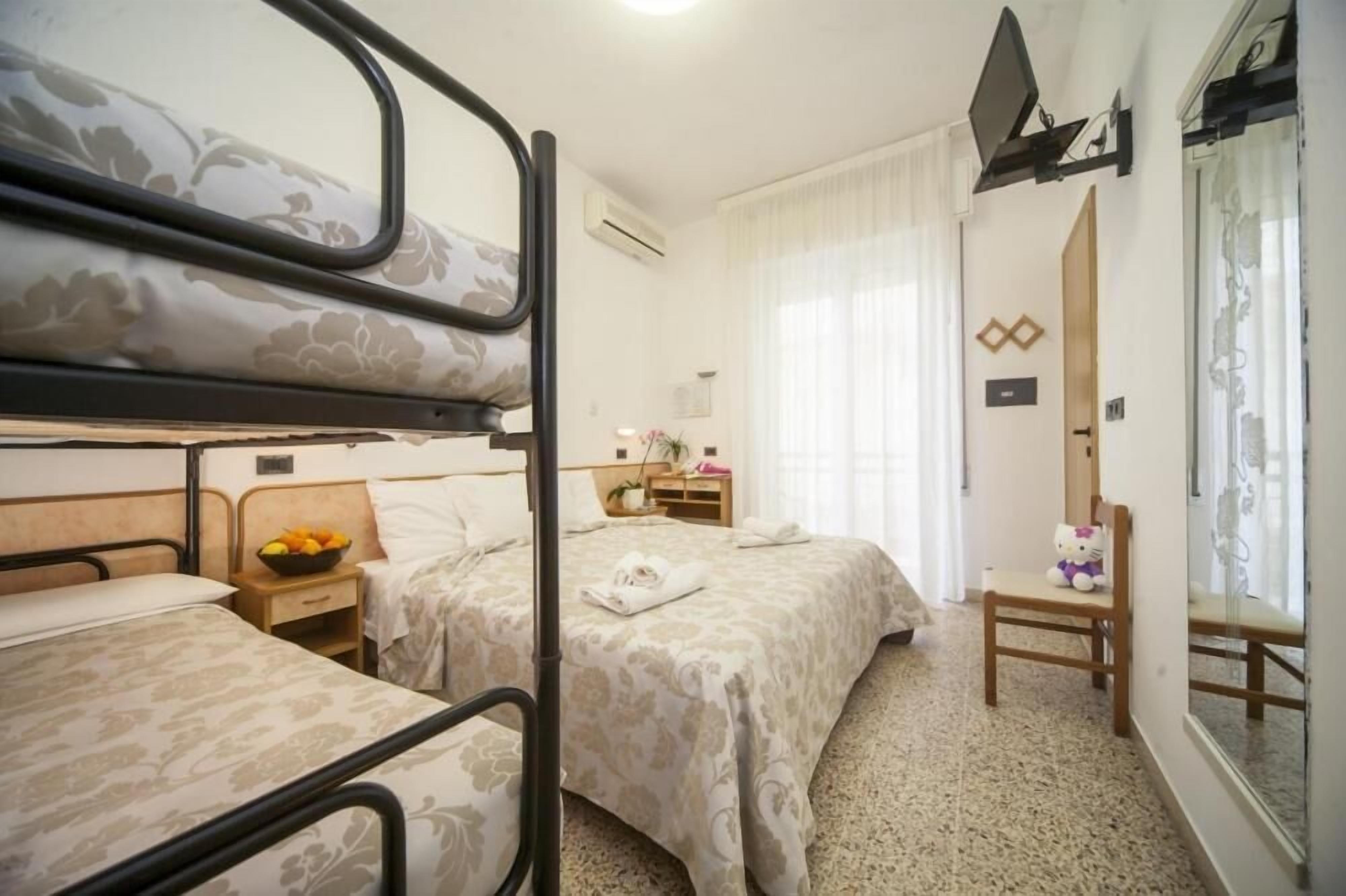 A hotel room with bunk beds, a bedside table with a fruit bowl, a chair, and a window with curtains.