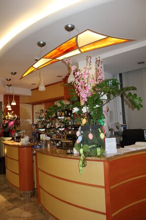 Zona de recepción de un hotel con un mostrador curvo, un arreglo floral grande y una barra provista de varias botellas.