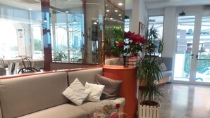 A modern hotel lobby with a beige sofa, a round coffee table with magazines, and a potted plant with red flowers. - Hotel Zurigo (Rimini)