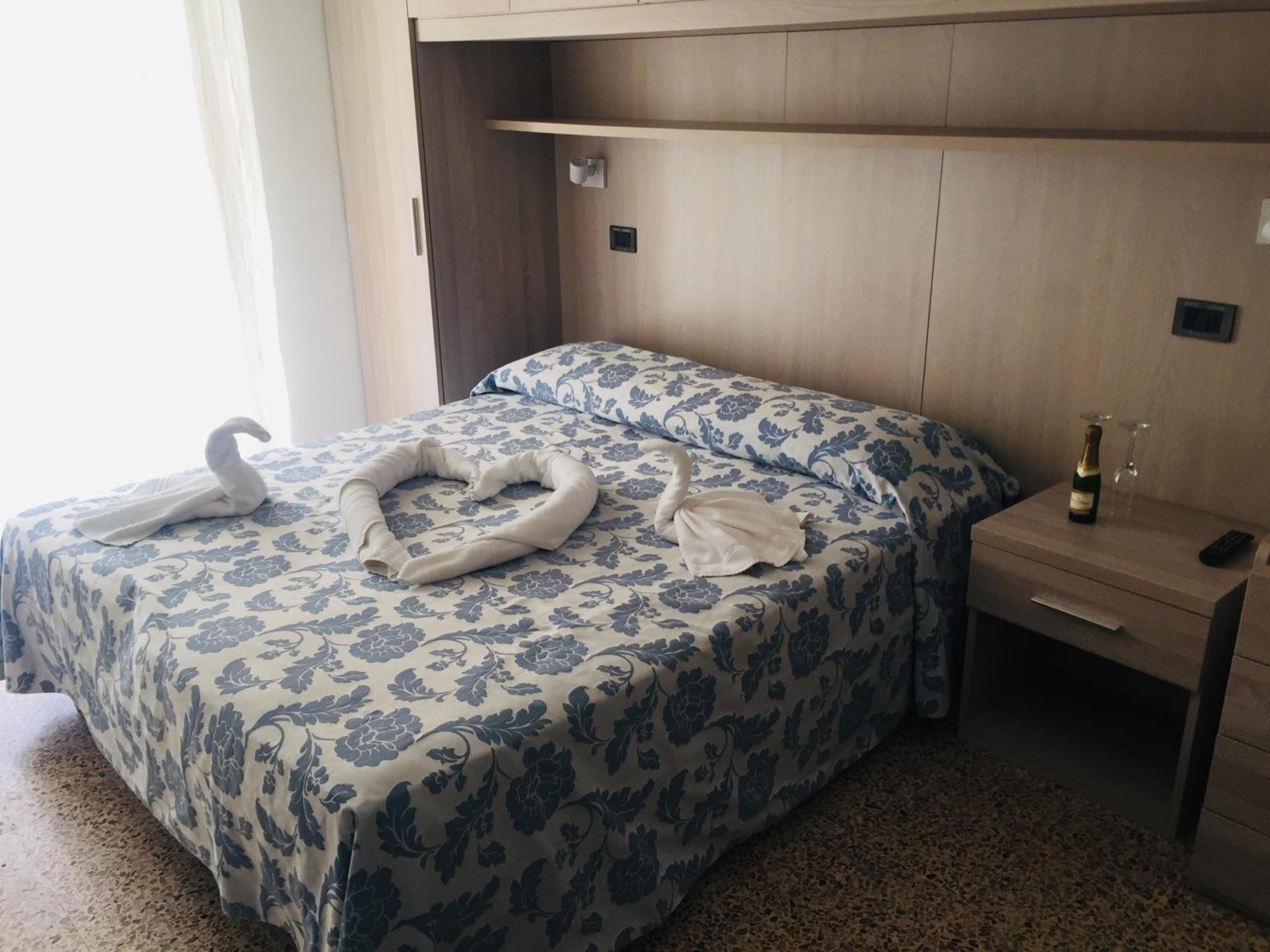 A hotel room with a bed featuring a floral bedspread, a nightstand with a bottle and glass, and a towel arrangement on the bed.