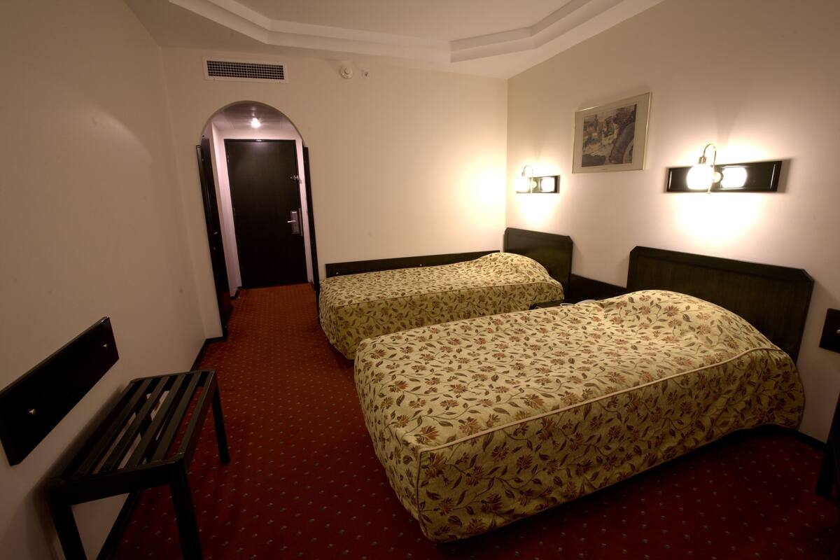 Habitación de hotel con dos camas, un banco, alfombra roja y una puerta que da a otra habitación.