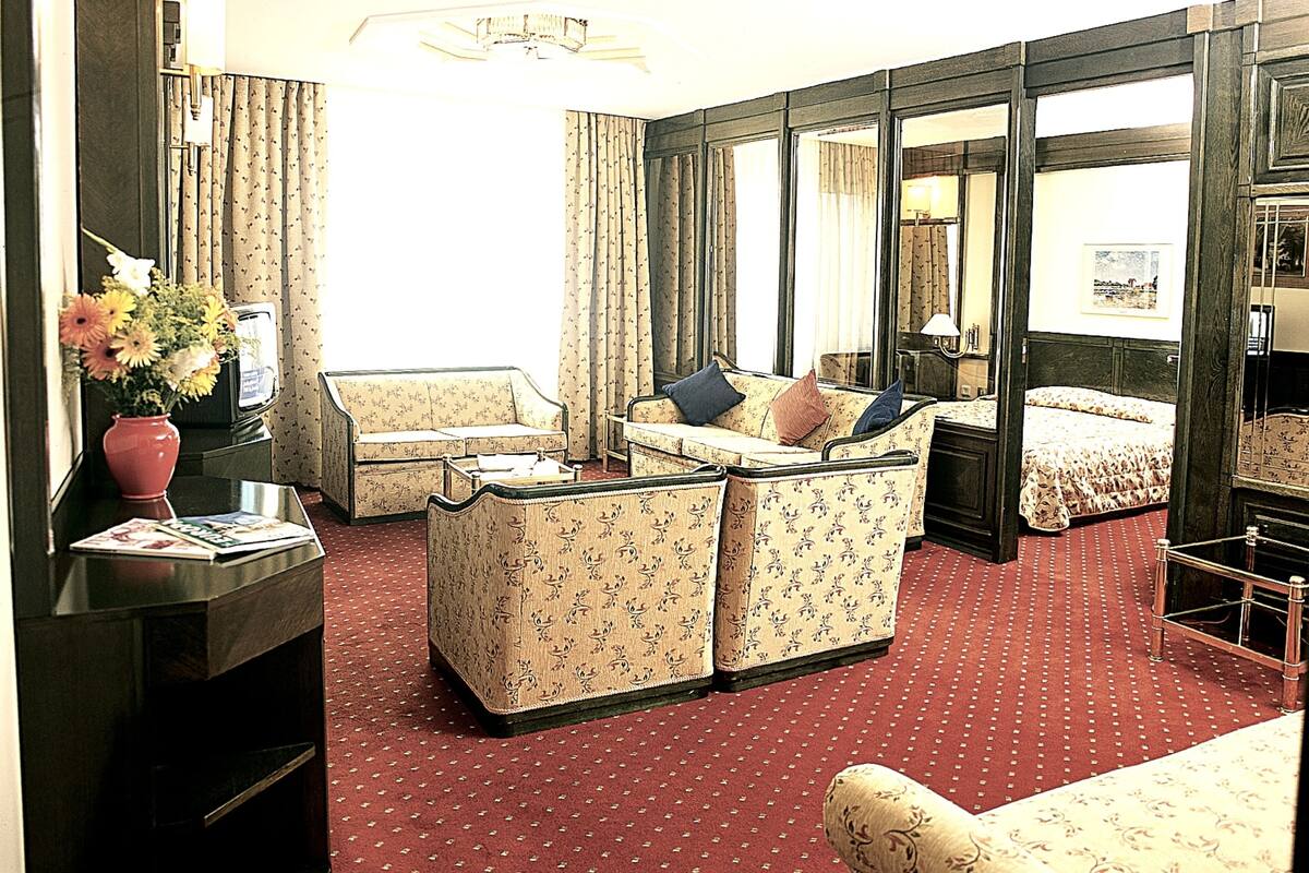 Habitación de hotel con sofá, una pequeña mesa con un florero, una cama con cubrecama estampado y un ventanal con cortinas.
