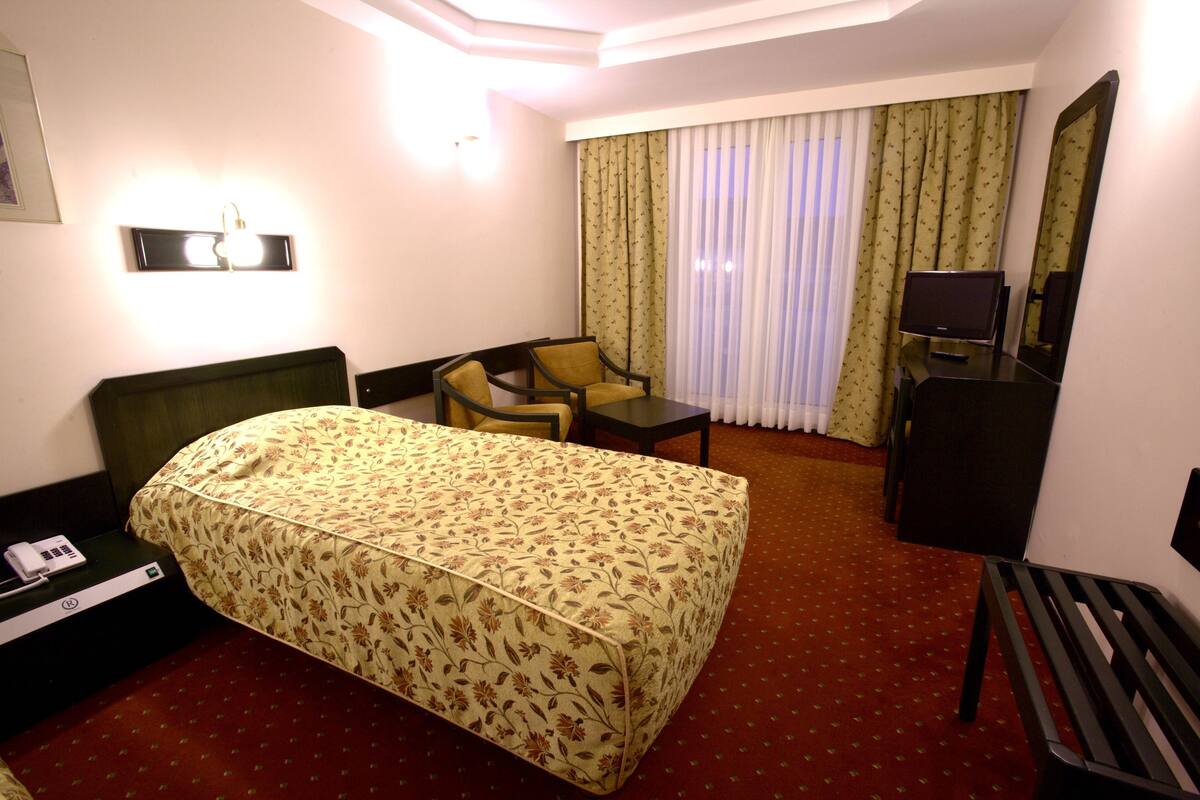 Habitación de hotel con cama, escritorio, silla, televisor y ventana con cortinas.