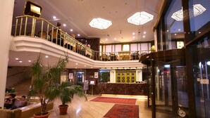 Un vestíbulo de hotel con mostrador de recepción de mármol, plantas en macetas, una escalera y un ascensor de vidrio.
