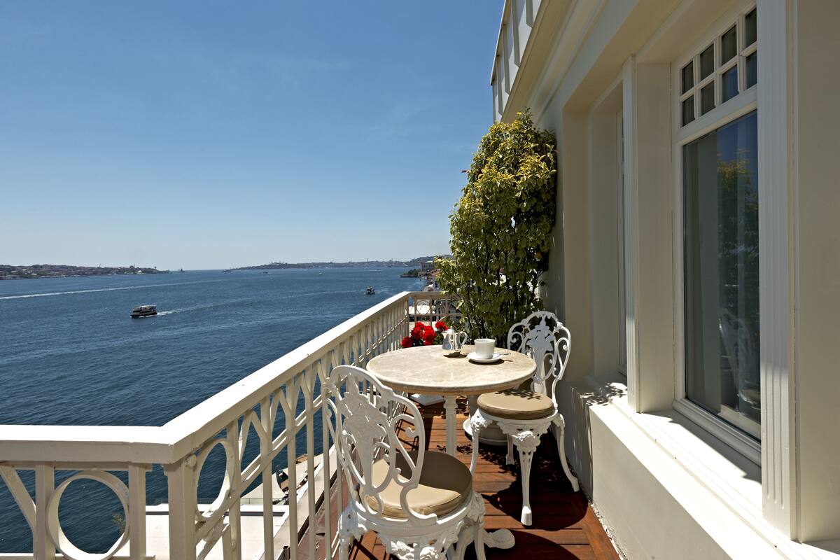A balcony with white ornate chairs and a table, overlooking a body of water with boats.