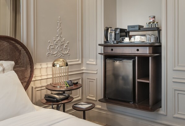 A hotel room with a bed, a bedside table with a lamp, a decorative wall piece, a minibar, and a small round table with a phone and a book.