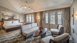 Ruime hotelkamer met een groot bed, een bank, een salontafel en een stoel. Er zijn meerdere ramen met gordijnen, een kroonluchter en een wandlamp.