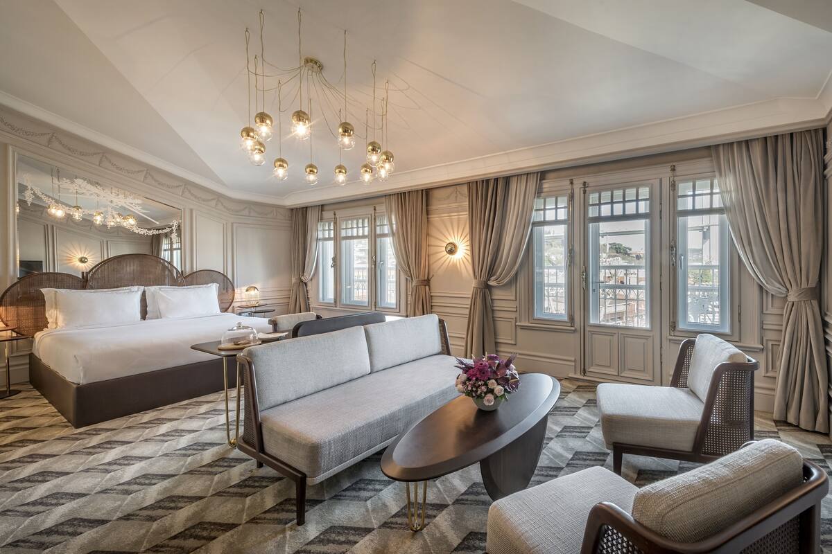 A spacious hotel room with a large bed, a sofa, a coffee table, and a chair. There are multiple windows with curtains, a chandelier, and a wall-mounted light fixture.