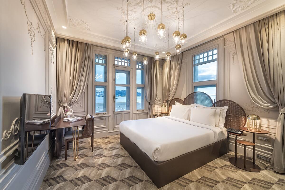 A spacious bedroom with a large bed, a desk, and a chair, featuring a patterned carpet and a chandelier.
