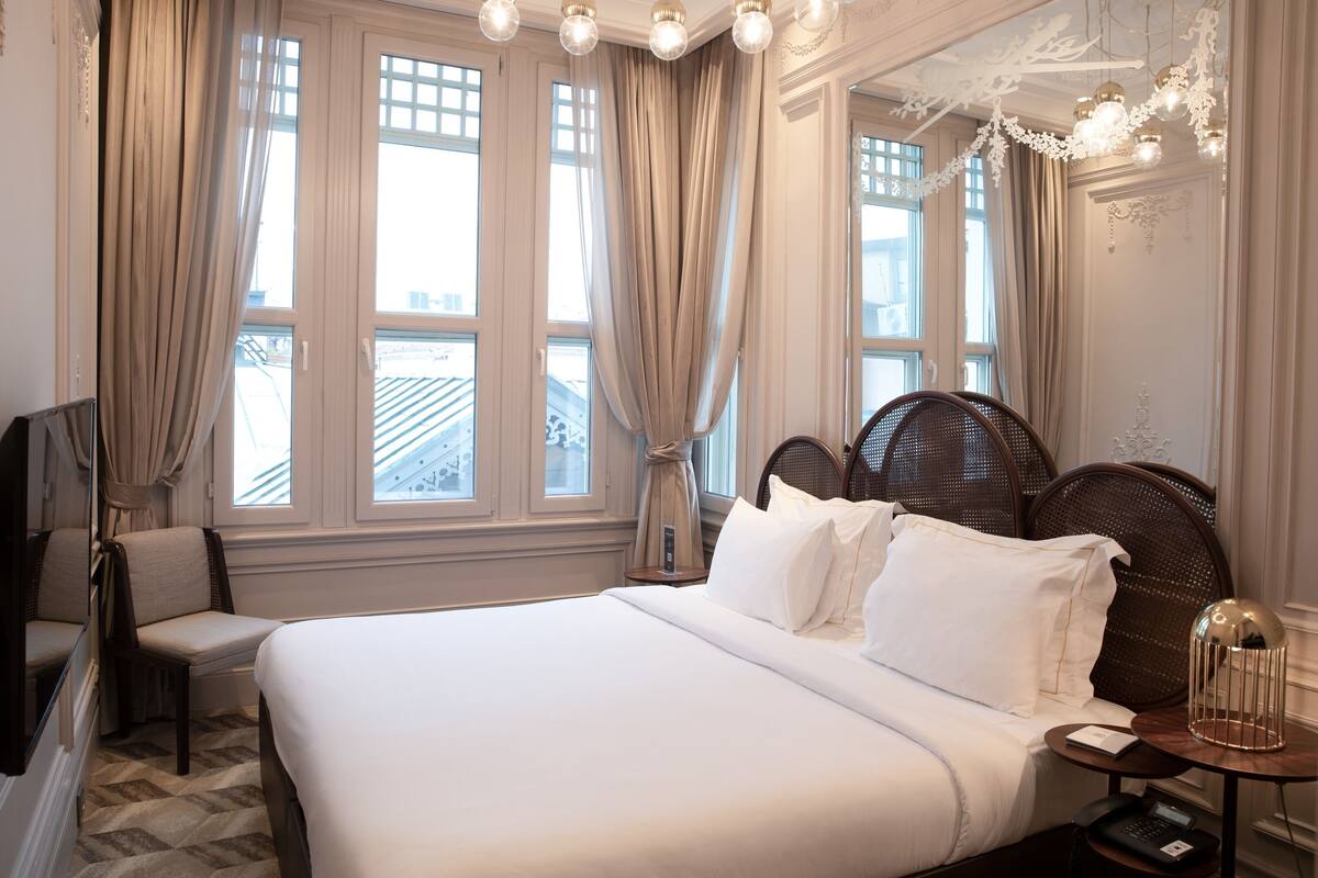 A hotel room with a large bed, a chair, a small table with a lamp, and a window with curtains.