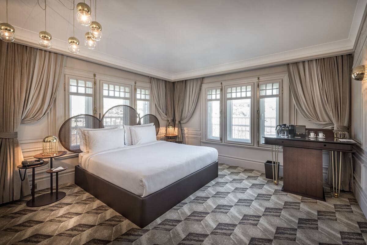A spacious bedroom with a large bed, a desk, and a chair. There are multiple windows with curtains, a chandelier, and a patterned carpet.