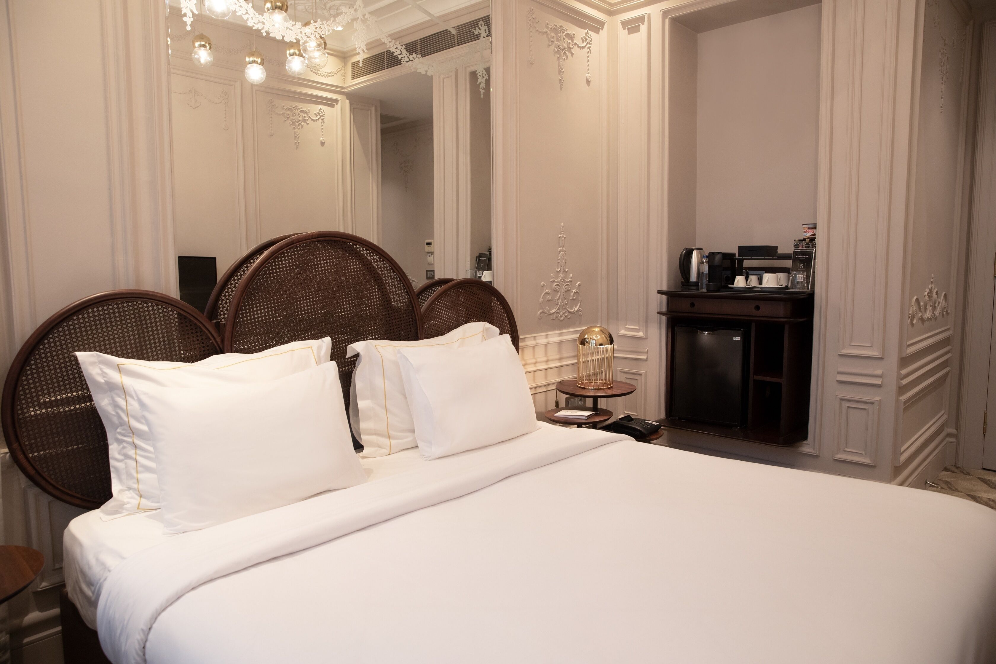 A neatly made bed with white linens and two large wicker headboards. A small bedside table with a lamp and a coffee maker is visible.