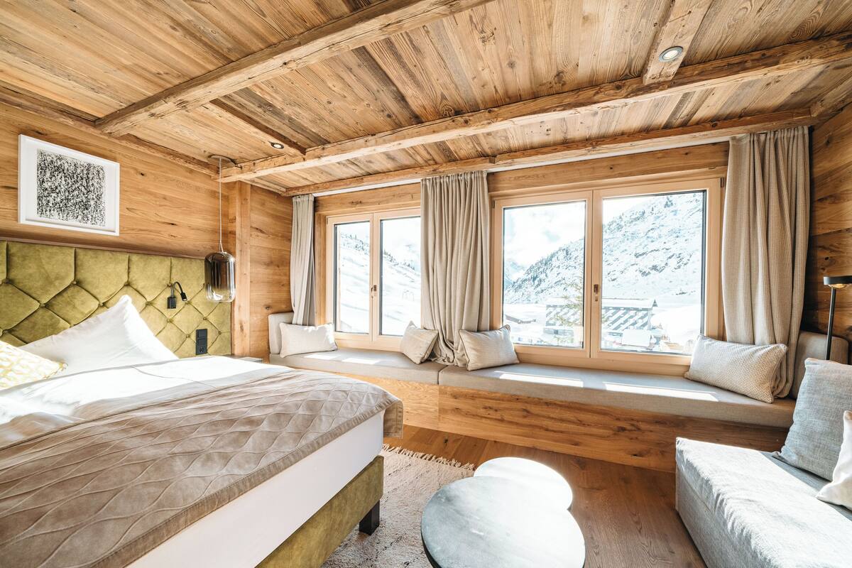 A wooden cabin bedroom with a bed, a sofa, and large windows overlooking a snowy mountain.