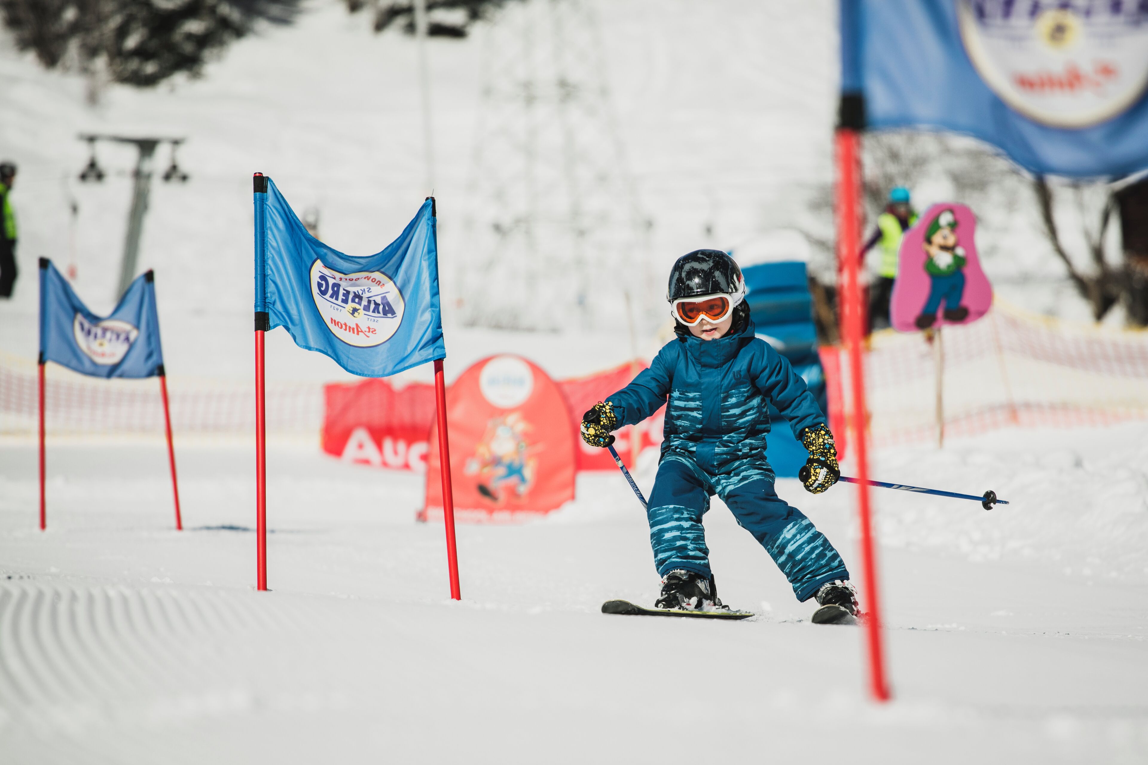 A child skiing on a snowy slope with flags and a red banner in the background.