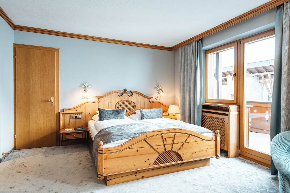A hotel room with a wooden bed, a nightstand, a lamp, a window with curtains, and a view of the outside.
