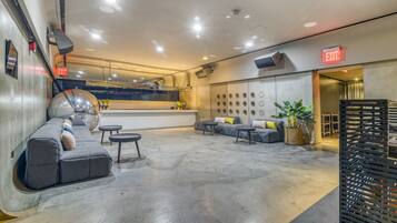 Un lobby moderno con suelo de concreto, sofás grises, un espejo redondo y un área de bar al fondo.