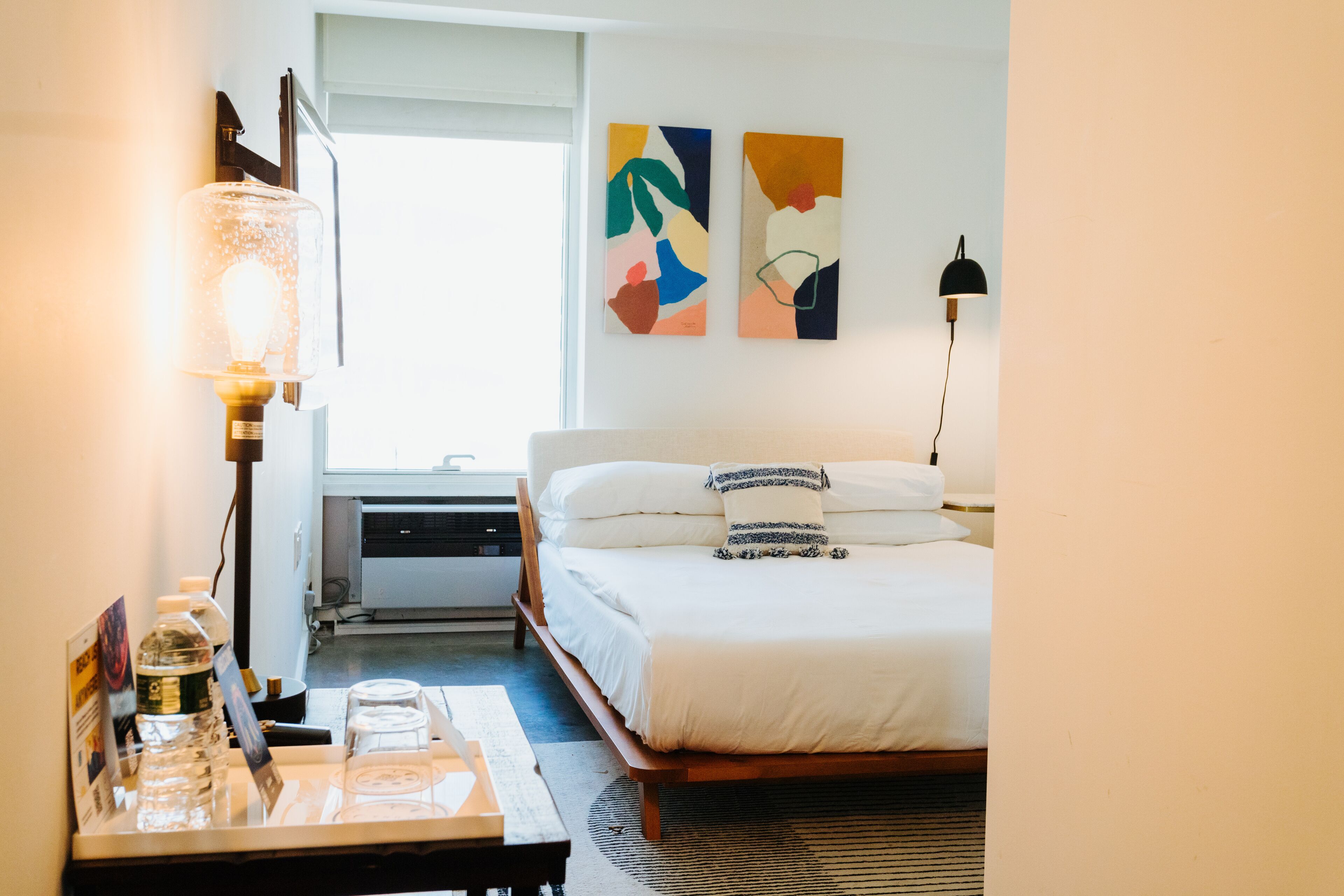 A hotel room with a bed, a desk with a lamp, a water bottle, and abstract art on the wall.