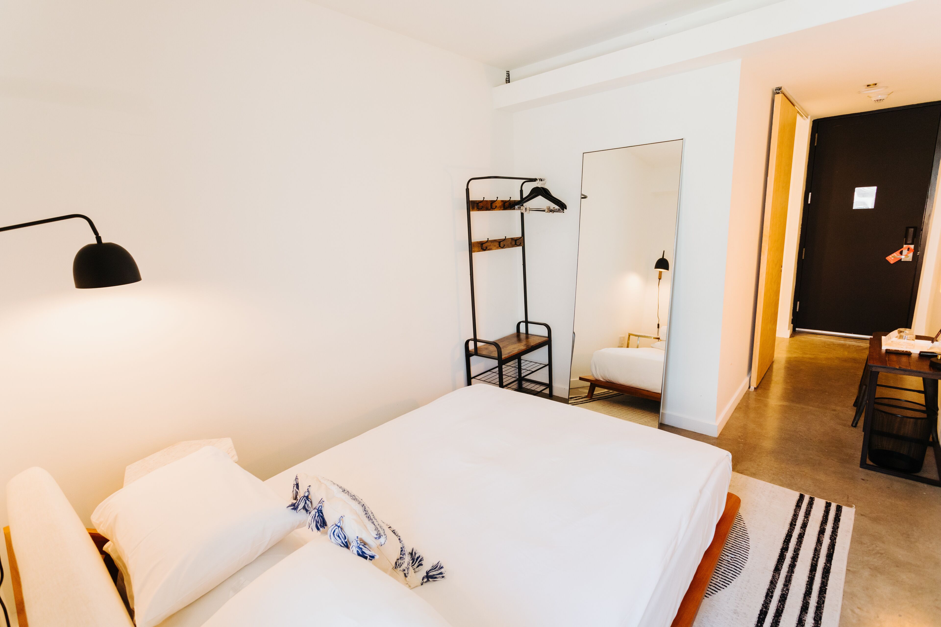 A hotel room with a large bed, a bedside table, a mirror, a coat rack, and a desk with a chair.