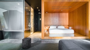 A modern bedroom with a wooden headboard, a bed with white bedding, a small stool, and a glass partition. - Hotel Alameda Highline (New York)