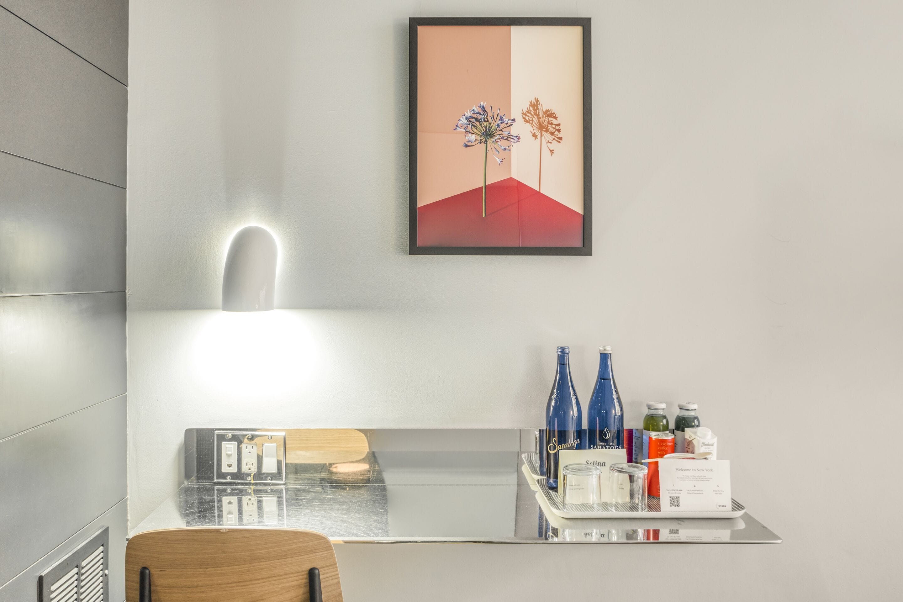 A modern hotel room with a minimalist design, featuring a desk with a computer, a chair, a framed artwork on the wall, and a tray with bottled water and a coffee machine.