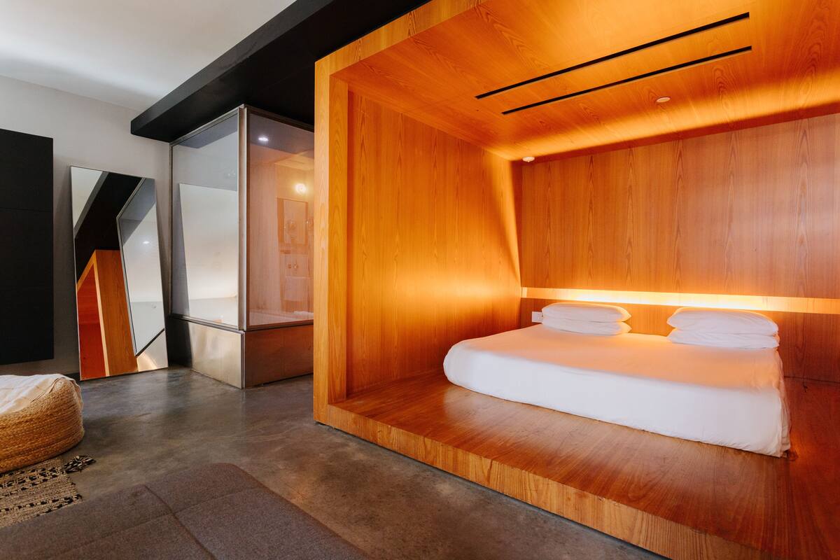 A modern hotel room with a wooden headboard, a bed with white linens, and a large mirror.