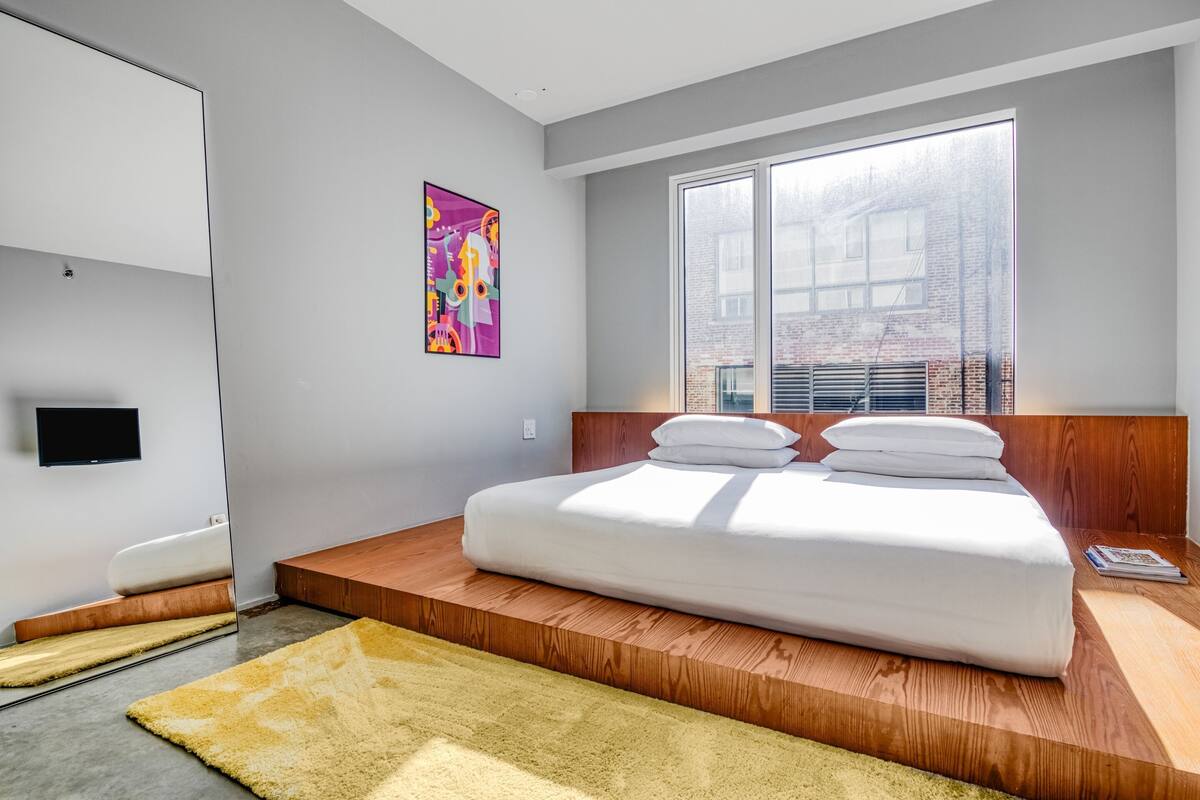 A modern bedroom with a large bed, a wooden platform, a large window, a flat-screen TV, and a colorful painting.