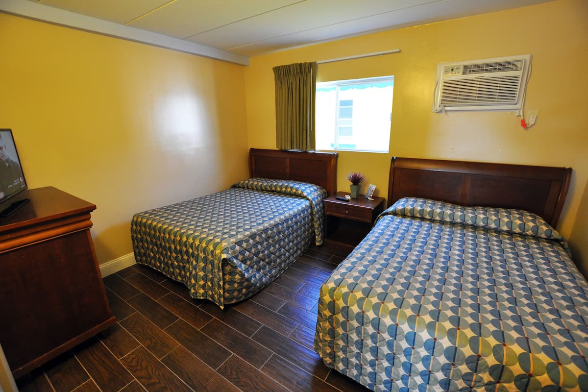 Une chambre d’hôtel avec deux lits, un plancher en bois, une table de chevet, une commode, une fenêtre avec des rideaux et un appareil de climatisation.