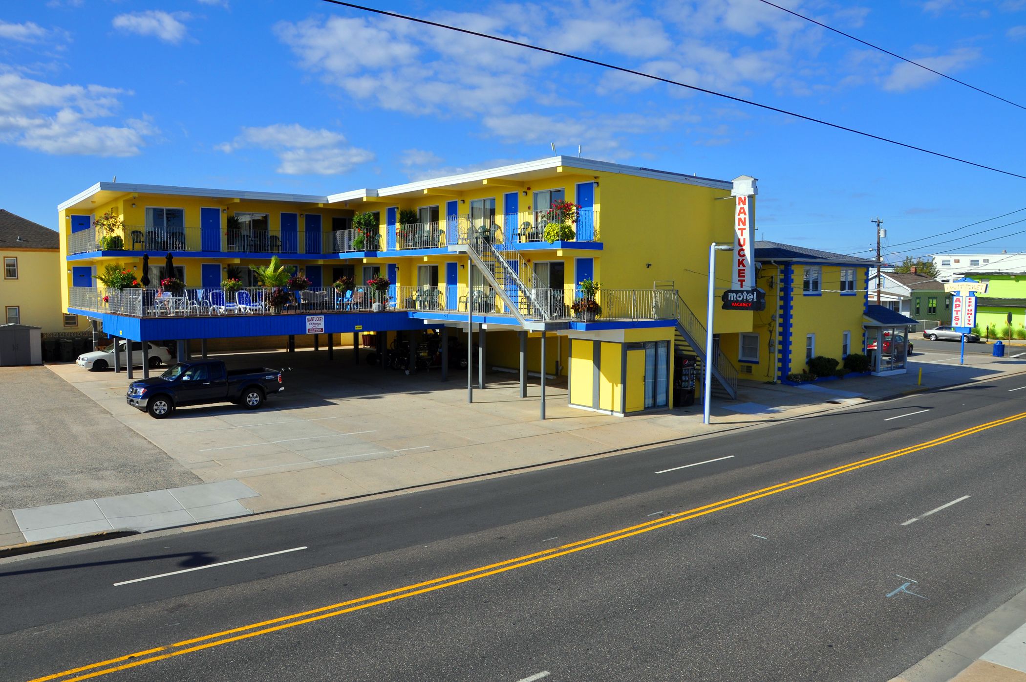 Un motel à la façade jaune et bleue, doté d’un parking couvert et d’un escalier bien visible menant à l’entrée.