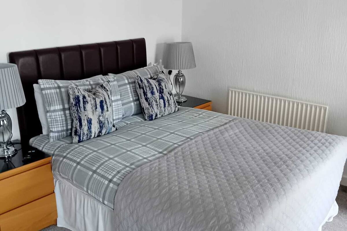 A neatly arranged bedroom with a large bed, two bedside tables, a nightstand, a lamp, a framed picture on the wall, and a radiator.