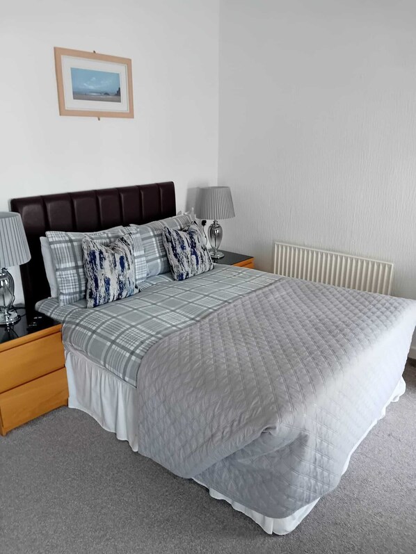 A neatly arranged bedroom with a large bed, two bedside tables, a nightstand, a lamp, a framed picture on the wall, and a radiator. - The Edelweiss (Blackpool)