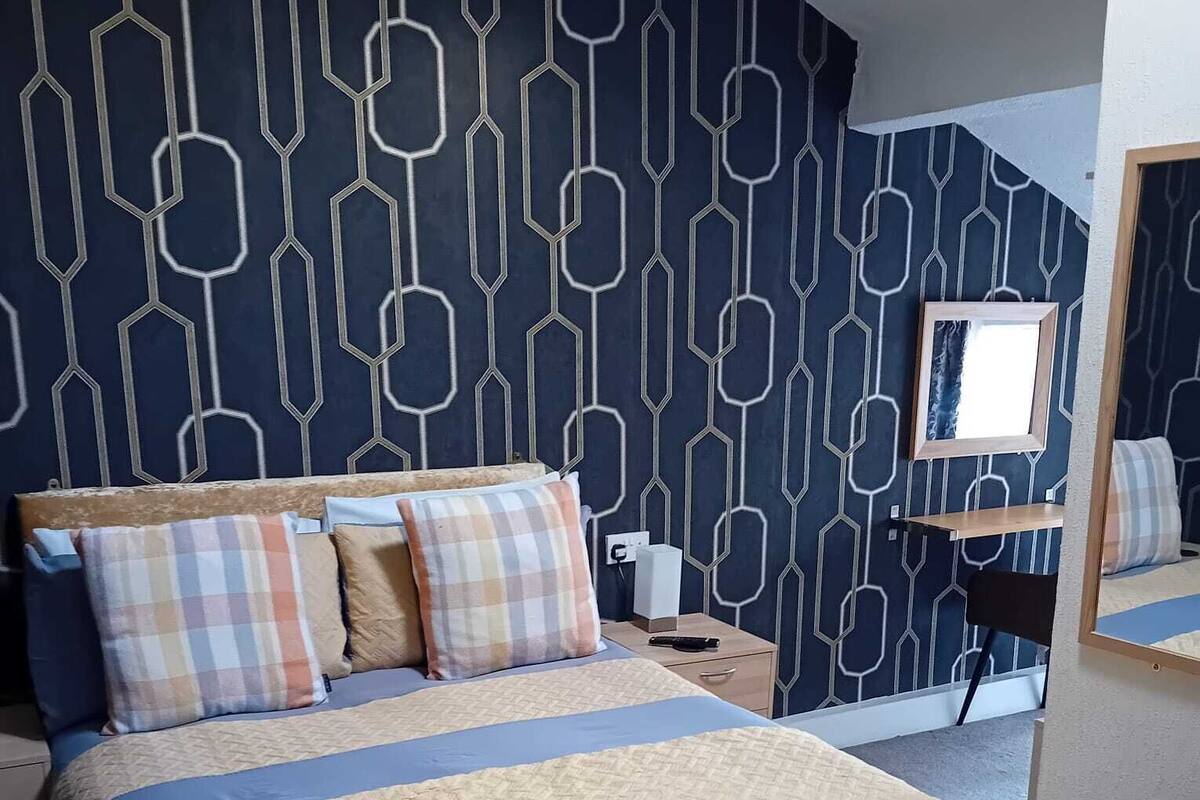 A bedroom with a bed, pillows, a nightstand, a mirror, and a wall with a geometric pattern.