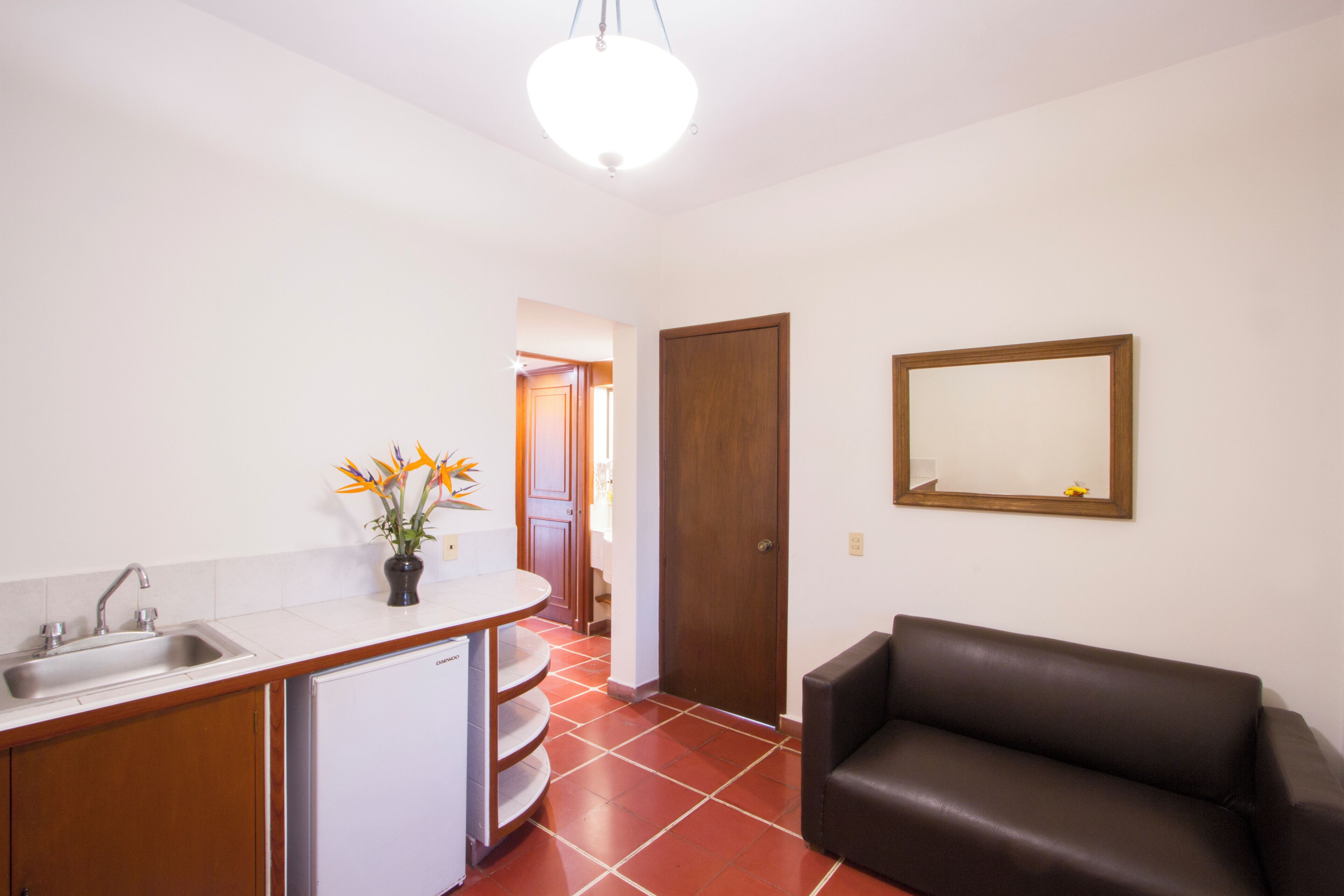 Una cocina compacta con un lavamanos, un refrigerador pequeño y un armario de madera. Hay un sofá de cuero marrón y un espejo en la pared.