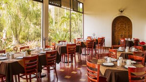 A restaurant with multiple tables and chairs, large windows with a view of greenery, and a wooden door with an arch. - Hotel Posada Loma (Fortin de las Flores)