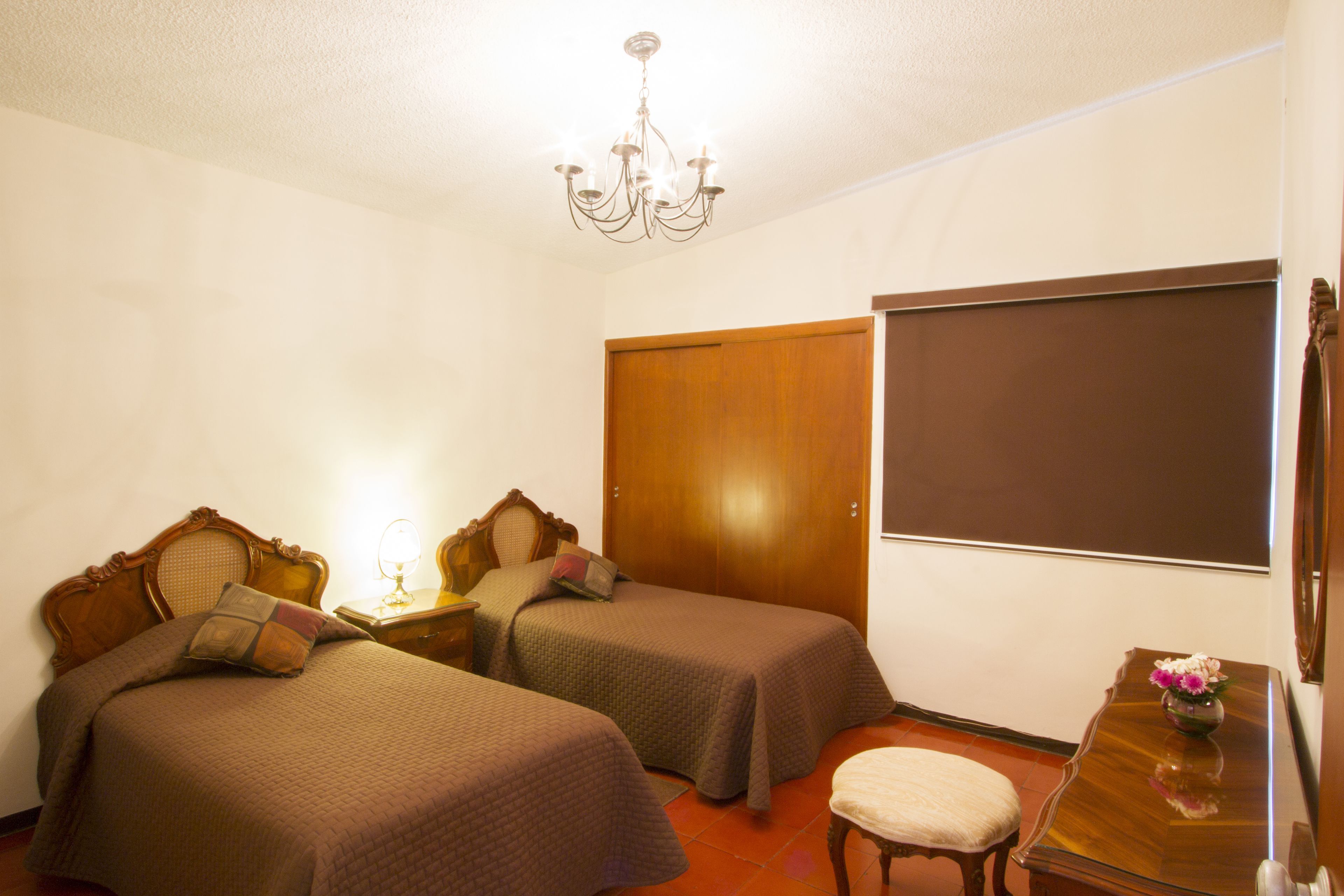 A bedroom with two single beds, a wooden wardrobe, a small table with a vase, and a chair.