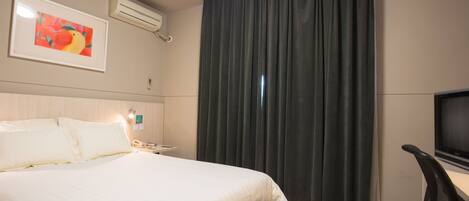 A hotel room with a bed, a desk with a chair, a television, and a large window with curtains.