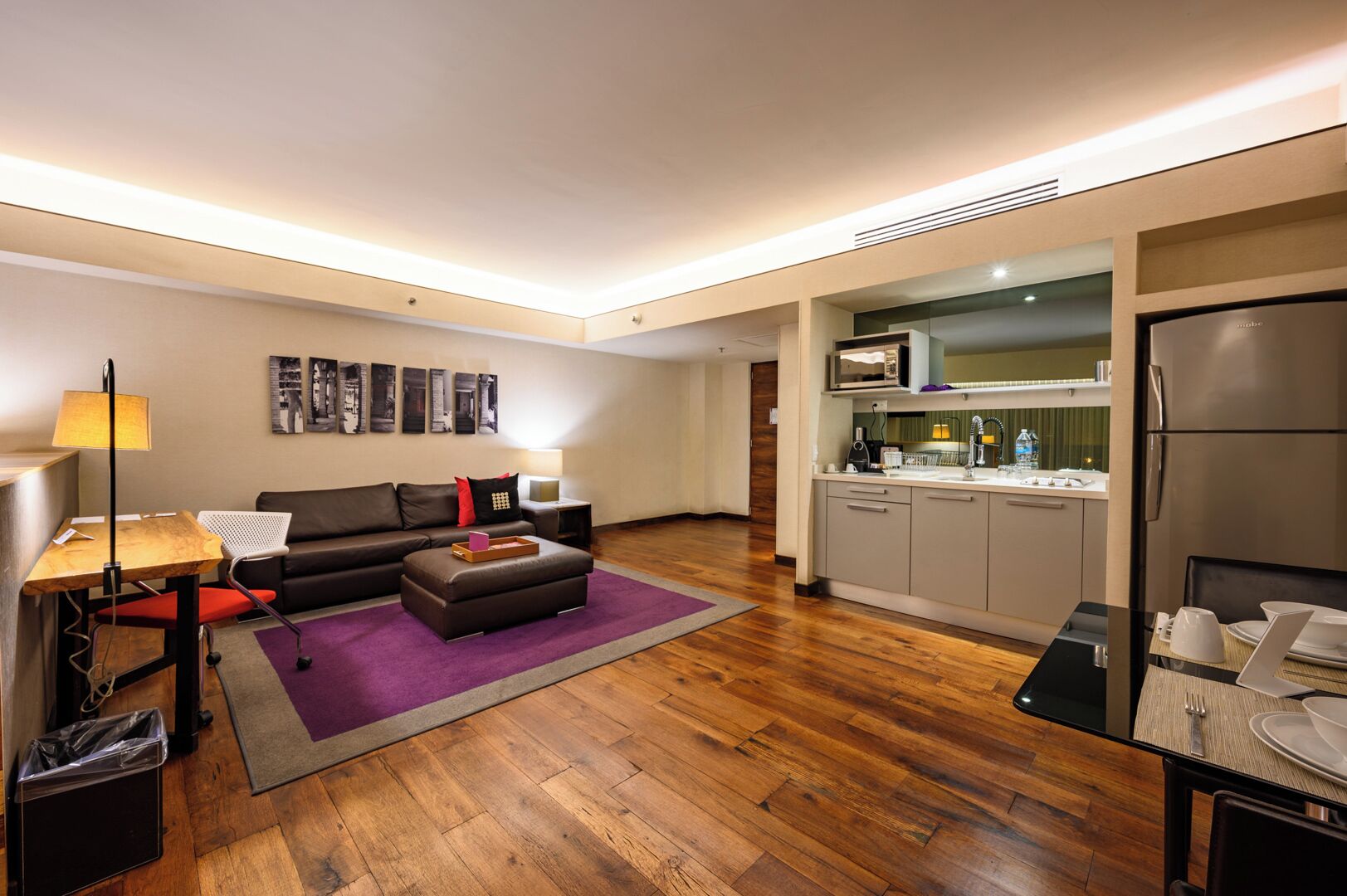 Un espacio habitable compacto con sofá, reposapiés y una pequeña mesa de comedor. Hay una kitchenette con microondas, fregadero y refrigerador. La habitación tiene suelo de madera y una alfombra morada.