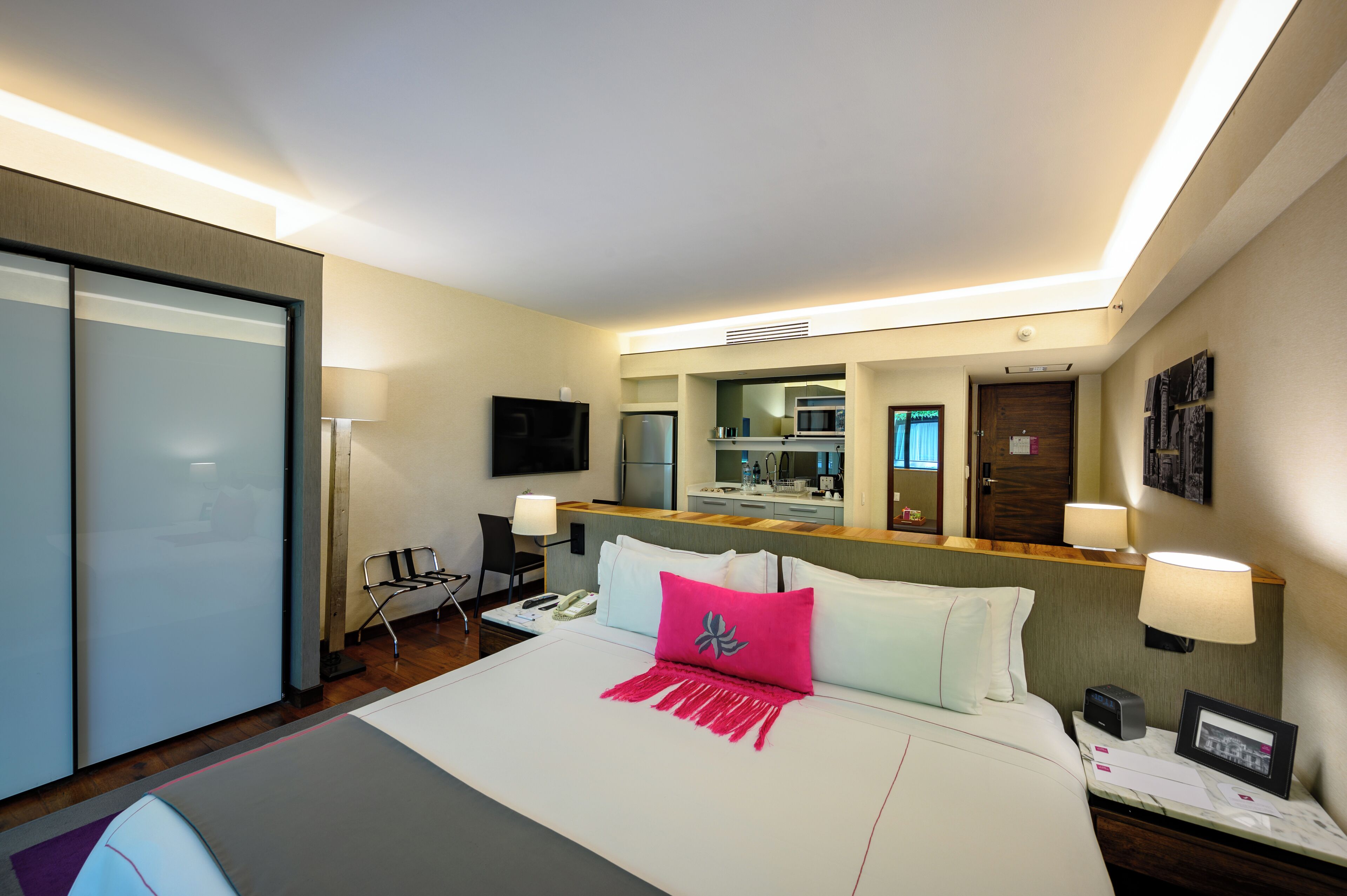 A hotel room with a bed, bedside table, lamp, and a view of the kitchen area.