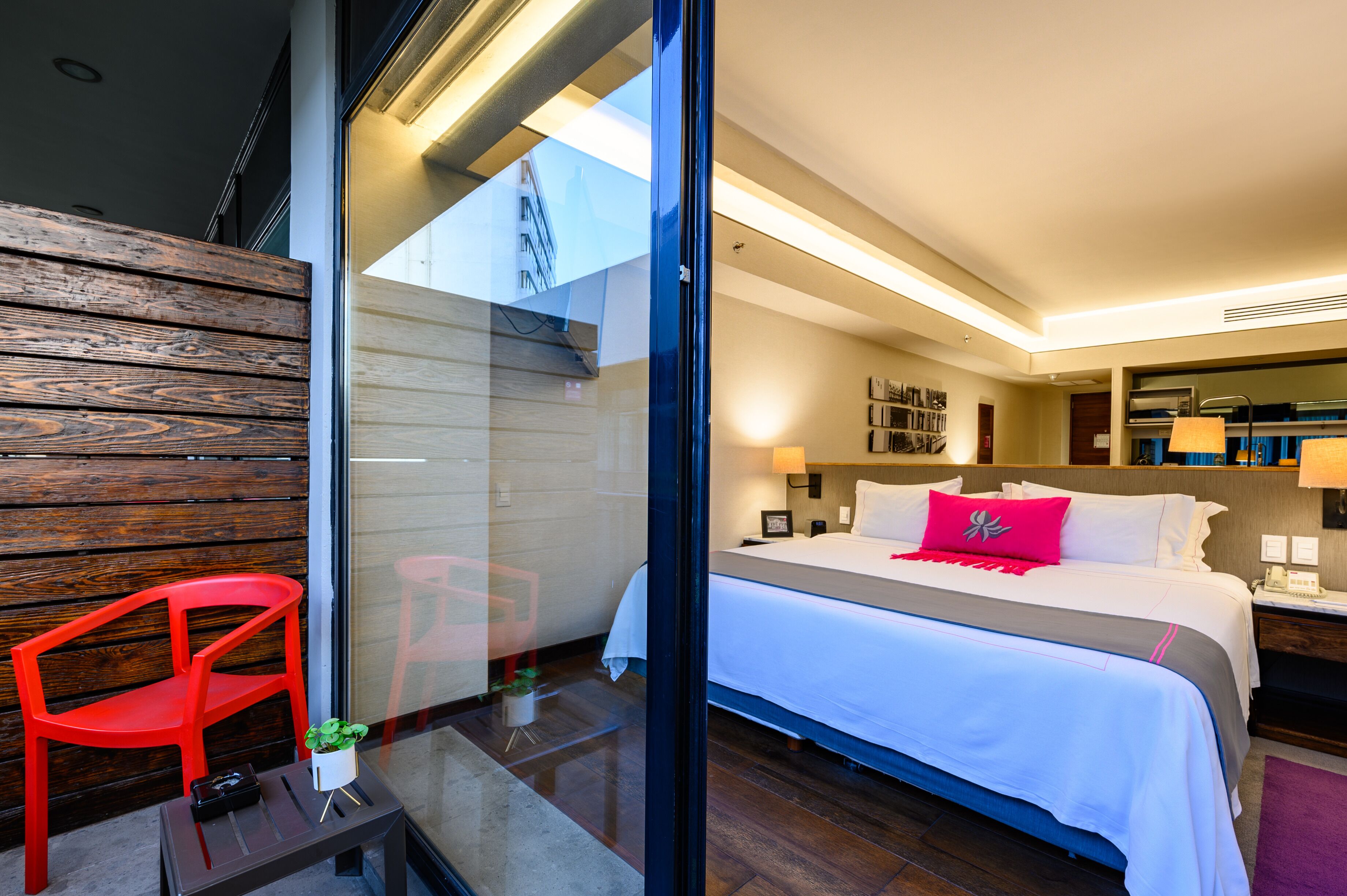 A hotel room with a large bed, a red chair, a small table with a plant, and a view of the outside through a glass wall.