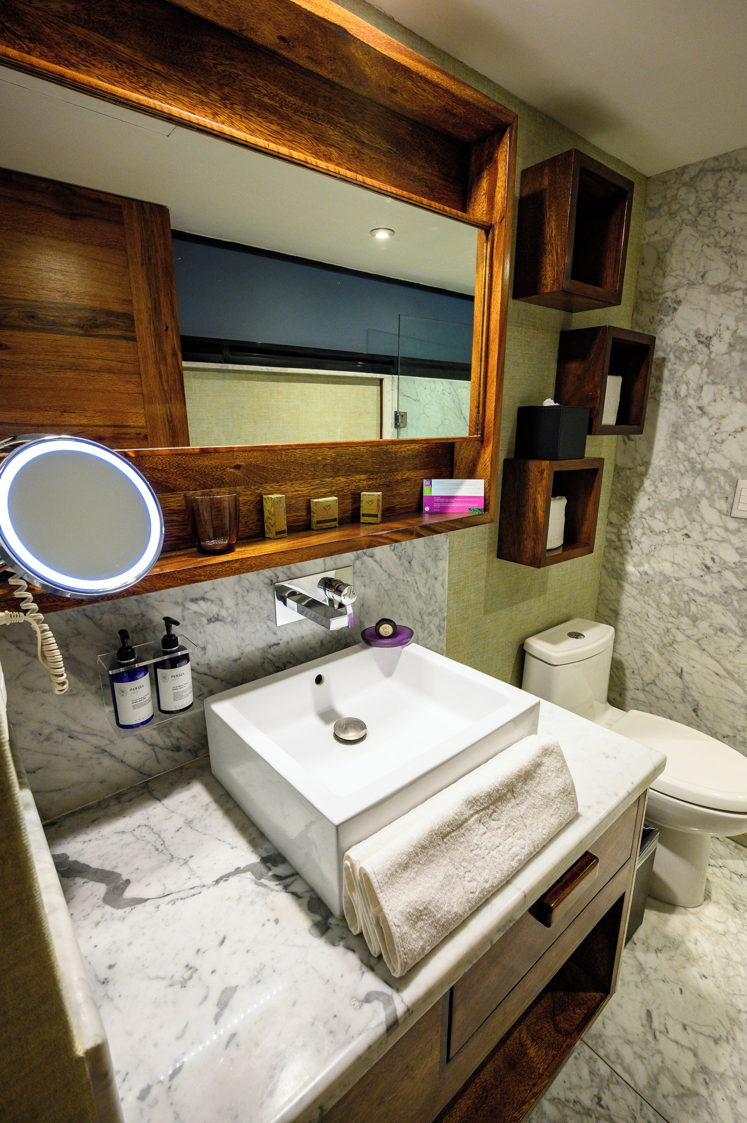 Een badkamer met een marmeren wastafel, een houten wastafelmeubel, een spiegel met verlichting, een toilet en een plank met toiletartikelen.